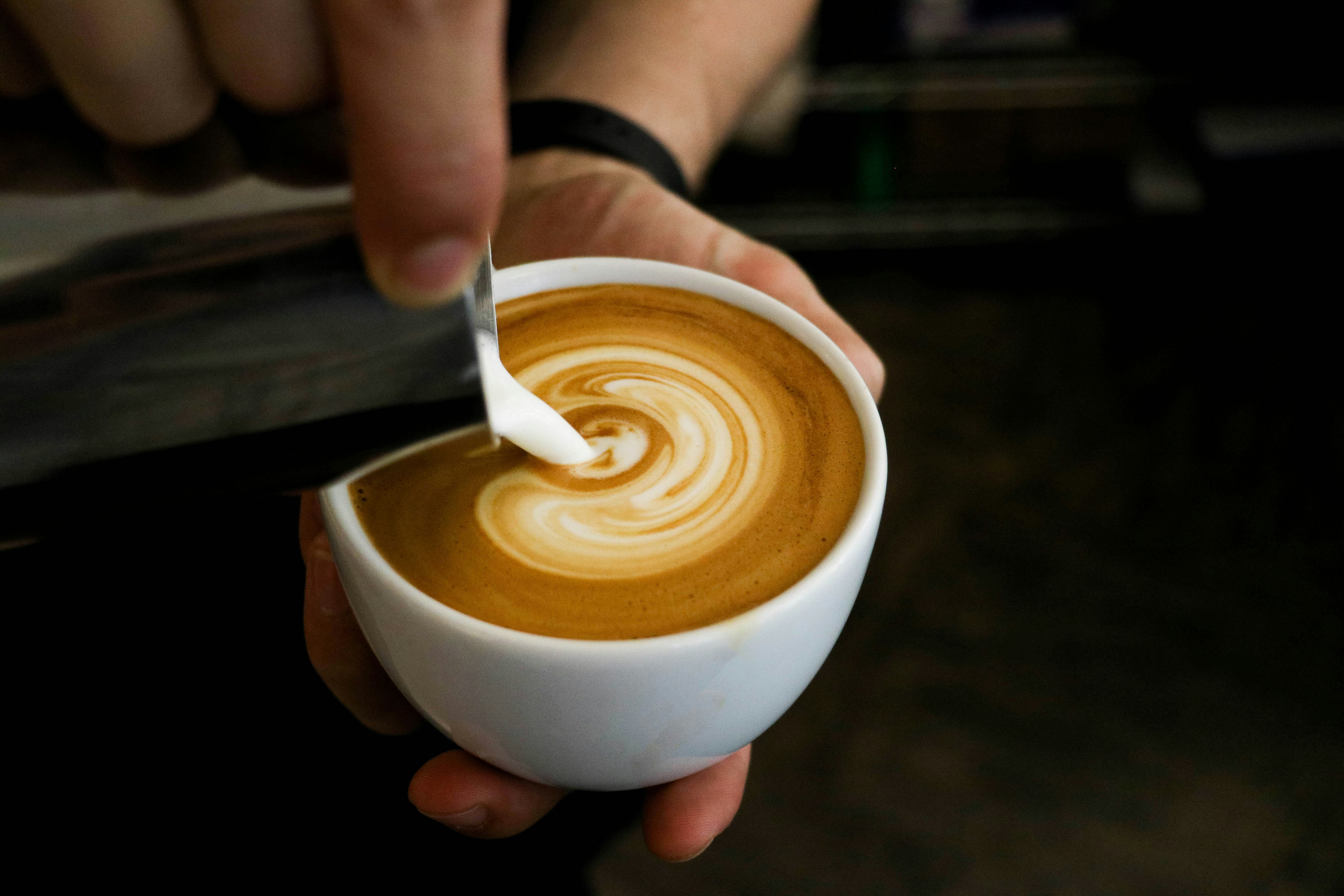 커피를 가득 채운 동그랗고 하얀 커피잔에 쇠로 된 주전자에 들어 있는 크림을 쏟아서 붓고 있는 사진