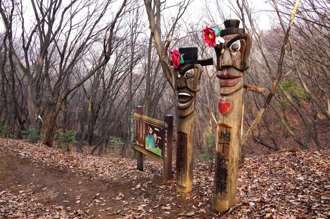 회심고개에 장승 2&#44; 천하대장군&#44; 천하여장군&#44; 그 옆에 회심고개 안내판&#44; 뒤로는 우거진 숲속&#44; 장승 머리 우측에 큰 붉은 꽃을 꽂았네요.