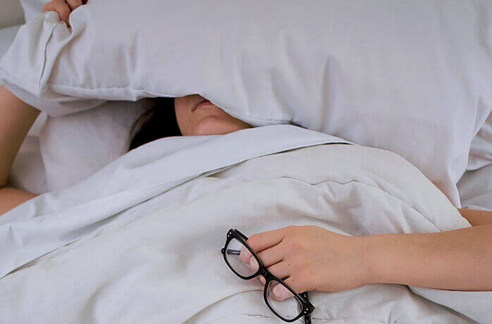 안경을 손에 쥔 채로 베개로 얼굴을 가리고 침대에 누워있는 한 여자의 모습