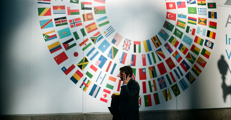 흰 벽에 세계 여러 나라의 국기가 그려져 있고 그 앞을 한 남자가 통화를 하며 지나가고 있다