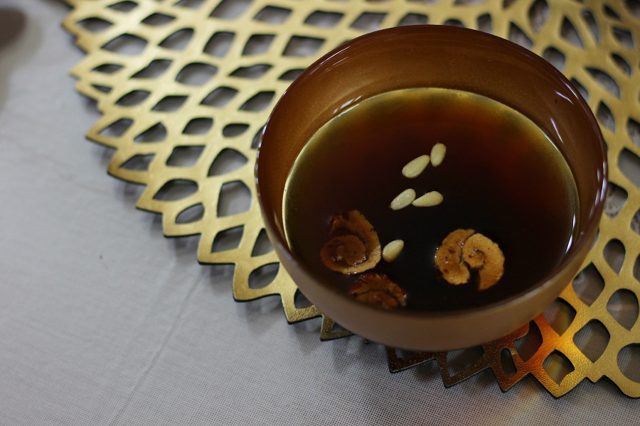 테이블 위에 금색의 깔판을 깔고 그 위에 잣과 대추를 편으로 썰어 놓은 수정과를 가득 담은 동그랗고 갈색의 그릇을 놓고 위에서 찍은 사진