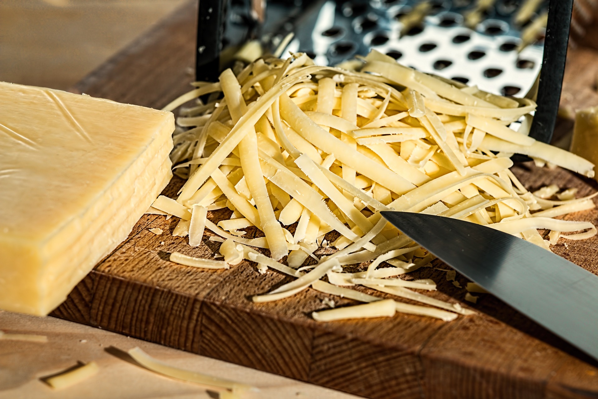 나무 테이블 위에 노란 치즈 덩어리를 두고 칼로 치즈를 반쯤 잘라놓고&#44; 그것을 편으로 잘라 수북하게 쌓아놓고 찍은 사진