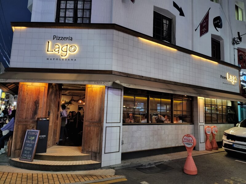 하얀 벽돌 간판에 Pizzeria Lago 라고 쓰여 있다. 오픈된 가게 안으로 노란색 조명이 새어나온다.