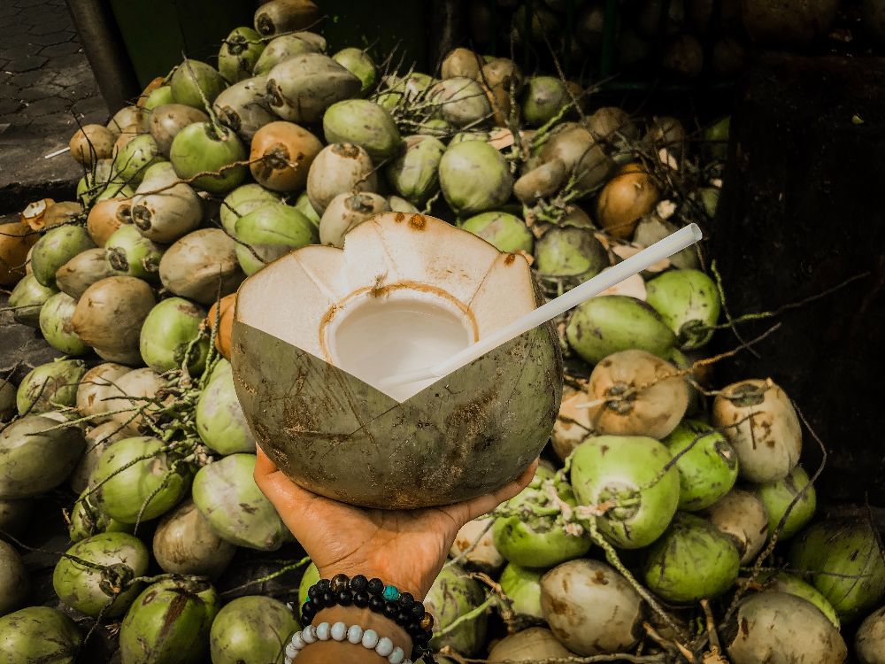 누군가가 빨대가 꽂힌 코코넛 열매를 팔찌를 낀 손으로 들고 있고&#44; 그 뒤에 코코넛이 가득 쌓여 있는 사진