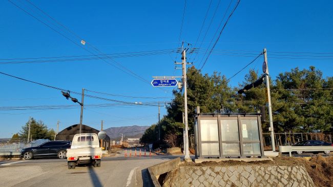 푸른 하늘&#44; 횡딘보도에 신호 댁중인 흰색 트럭 1&#44; ㄱㄹ 건너 숲&#44; 왼쪽으로 지나가는 검은 승용차 1&#44; 뒷 모습이 보이는 버스 정류장&#44;