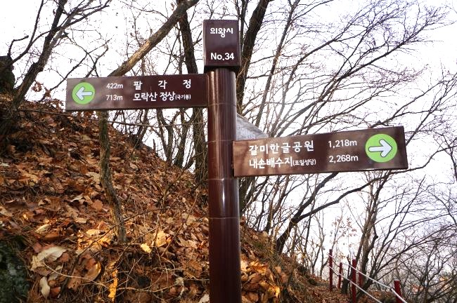 이정표&#44; 모락산 정상까지 713m&#44; 진한 고구마색에 흰글씨&#44; 화살표는 둥근 녹색 원 안에 흰색&#44; 뒤로는 잡목&#44; 하얀 하늘&#44;