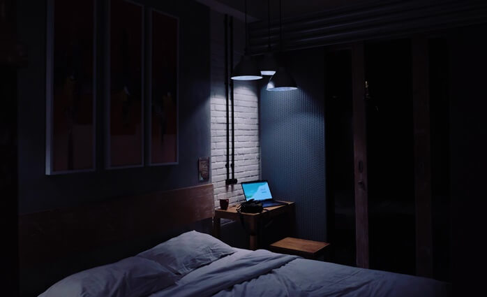 어두운 침실 안, 침대 옆에 켜져있는 노트북에서 밝은 빛이 나오고 있는 모습