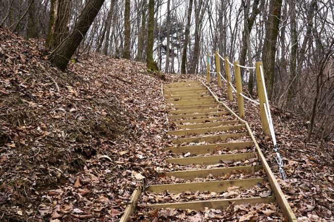 오르막 급경사 나무계단&#44; 우측에 안전로프 난간 두 줄&#44; 왼쪽 낙엽 수북한 산비탈&#44; 뒤로는 우거진 숲&#44;