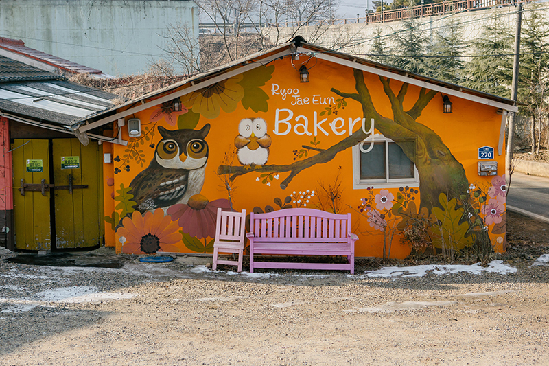 류재은 베이커리 물류창고. 오렌지색 벽에 부엉이 그림이 그려져 있다. 건물 앞에는 핑크색 의자와 벤치가 놓여있다.