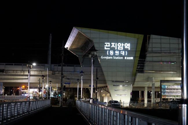 어두운 하늘&#44; 회색 건물벽의 곤지암역&#44; 흰색 LED 큰 글씨&#44; 아래는 건지암천 건너가는 보행교&#44; 양쪽에 안전 난간&#44;