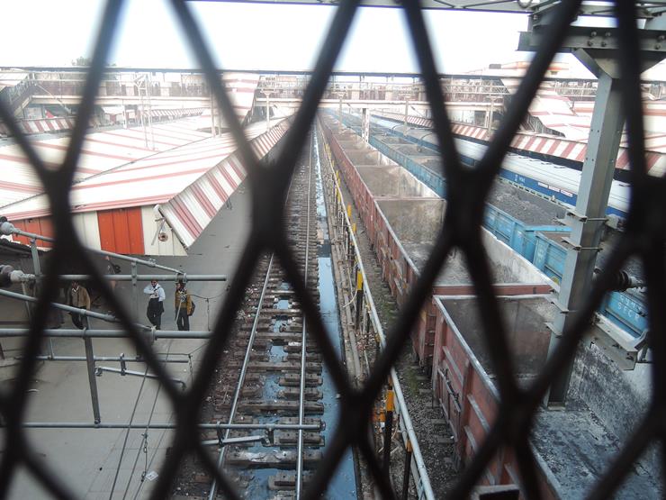 창살 밖으로 인도 화물 열차 두대와 플랫폼에 서 있는 사람들이 보이는 사진