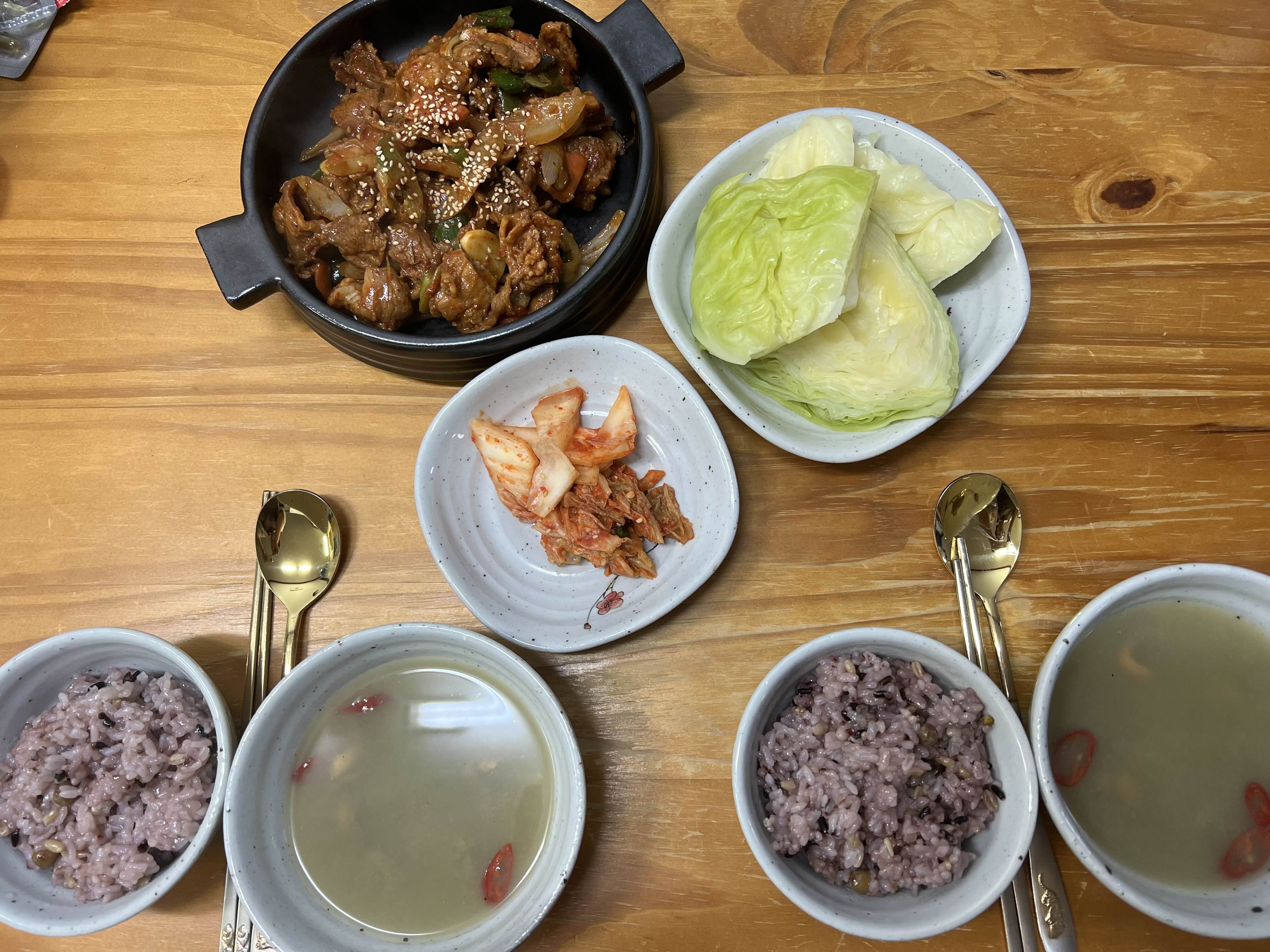 식탁 위에 돼지불고기, 양배추 쌈, 밥, 홍합탕, 김치가 놓여져 있는 사진