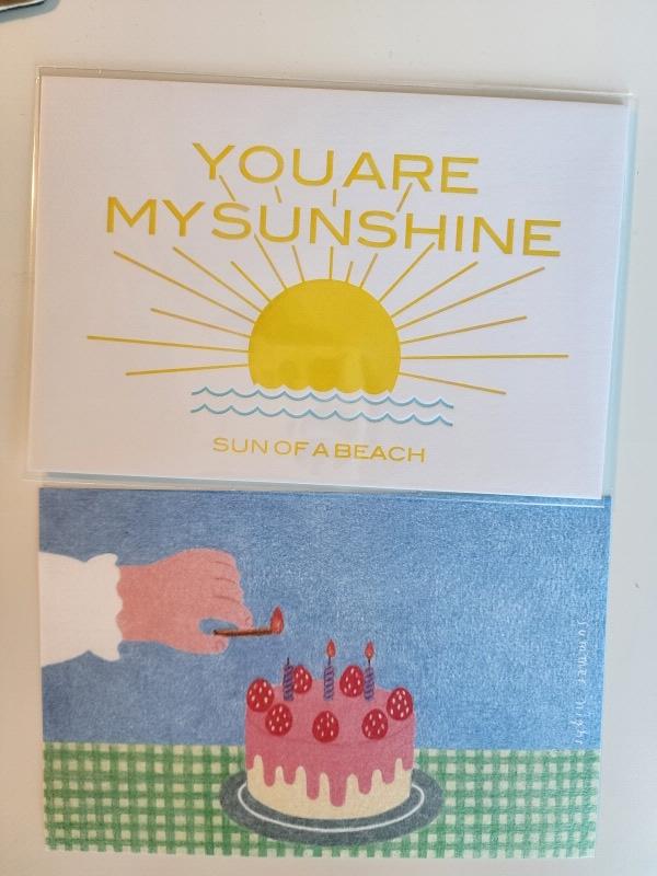 포셋에서 구매한 엽서들. 윗 쪽 엽서는 하얀 바탕에 일출하는 해 일러스트가 있고 &#39;YOU ARE MY SUNSHINE&#44; SON OF BEACH&#39; 라는 장난스러운 문구가 쓰여 있다. 밑에 엽서는 딸기 케이크에 성냥으로 불을 붙이는 소녀의 손이 프린트되어 있다.