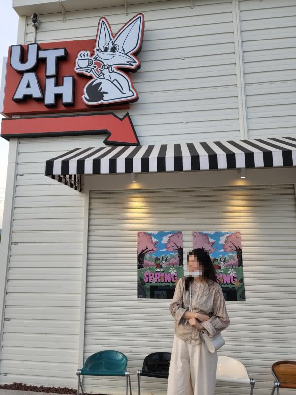 UTAH 라는 글자와 함께 커피를 든 여우 마스코트가 있는 유타 커피의 간판. 밑에는 검정색과 화이트 스트라이프로 된 차양이 있고 아래는 벚꽃 그림의 포스터가 붙여져 있다. 그 앞에서 여자가 포즈를 취하고 있다.