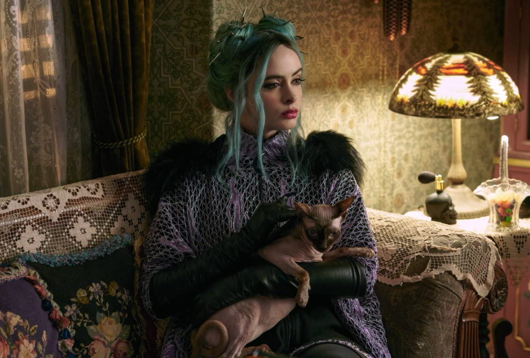 녹색 머리에 보라색 옷을 입고 가죽장갑을 낀 마녀 나타샤가 고양이를 가지고 쇼파에 앉아있는 장면 