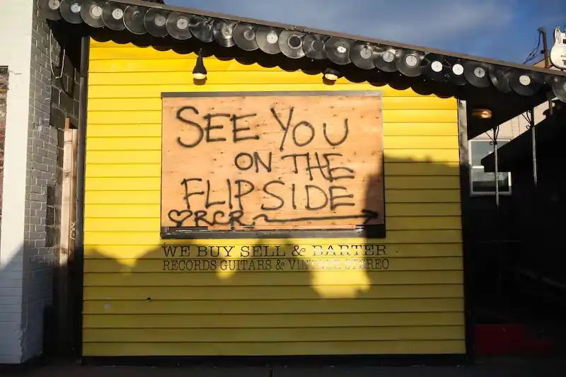 노란색 셔터가 닫힌 차고에 붙은 나무보드에 See you on the flipside라는 문구가 스프레이로 쓰여있다.