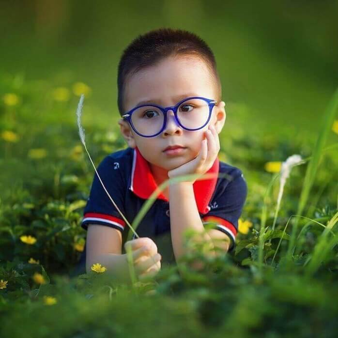 파란색 안경테를 쓴 어린 소년이 꽃밭 가운데에서 한 손으로 턱 괴고 앉아있는 모습