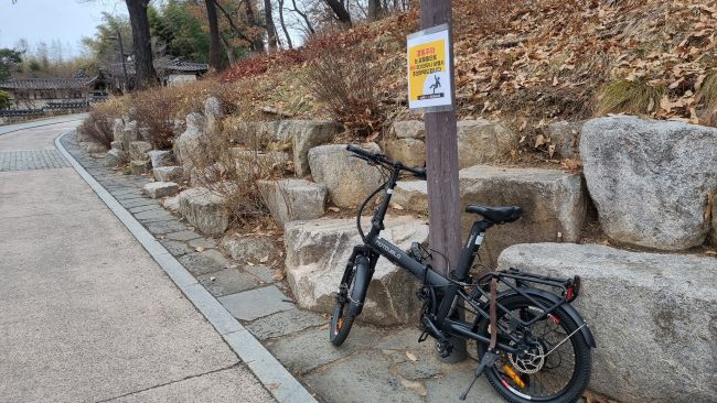 공원 보안등에 매어놓은 자전거&#44; 뒤로는 바윗돌들을 쌓아놓은 언덕 비탈면&#44; 왼쪽에 시멘트포장길&#44;