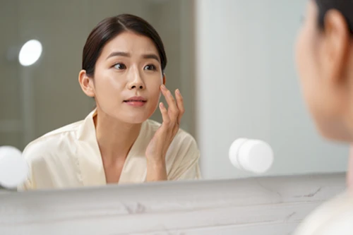 거울을 보며 개선된 피부결에 만족하는 한국 여성의 실사 근접 사진입니다.
