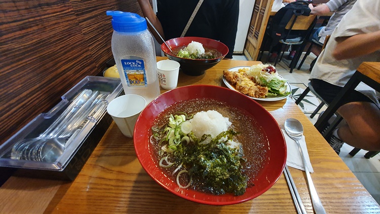 경성모밀 상차림 상 위에 냉모밀 살얼음 육수에 갈은 무 김가루 다진파 고명 돈가스 샐러드가 있다. 파란색 물병