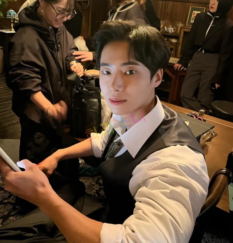 클래식한 실내 공간에서 흰 셔츠와 넥타이 위에 검은색 조끼를 입은 추영우가 휴대폰을 들고 정면을 응시하는 모습. 그는 부드러운 미소를 띠며 카메라를 응시하고 있으며&amp;#44; 주변에는 촬영 스태프들이 분주히 움직이고 있다.