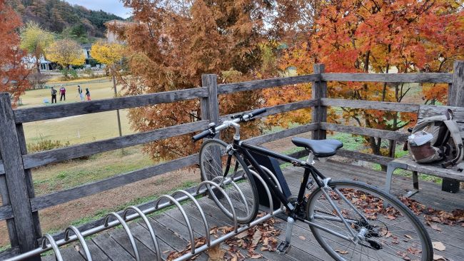 쉼터에 자전거&#44; 벤치 위에 내려 놓은 배낭&#44; 단풍나무 사이로 멀리 작게 보이는 파크골프 치는 네 사람&#44;