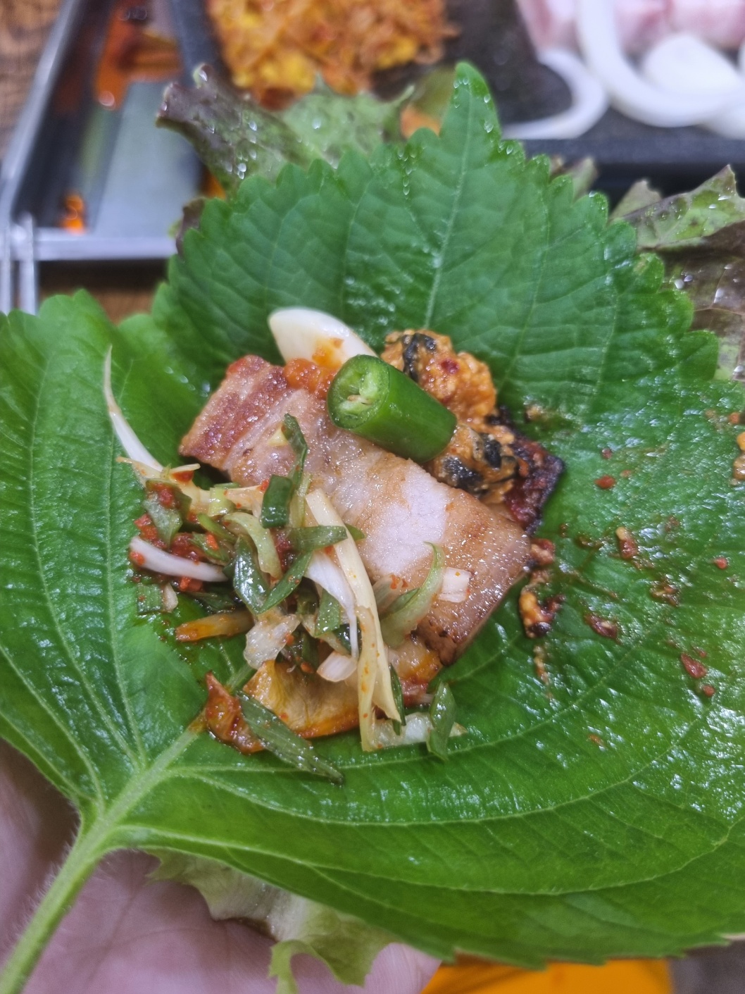 상추와 깻잎 위에 삼겹살&#44; 파채&#44; 우렁강된장&#44; 고추&#44; 마늘이 올려져 있다.