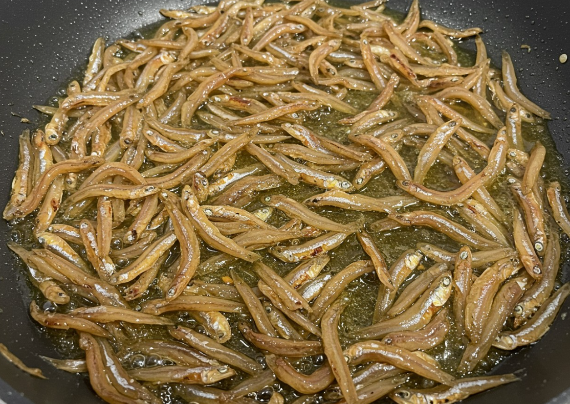 윤기 나는 양념이 멸치 전체에 고르게 퍼지는 모습. 프라이팬 가장자리에는 은은한 기름 코팅이 보임.