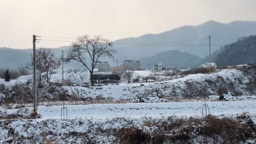 눈 덮인 들판 마을풍경, 뒤로 야산, 흐린 하늘, 좌우 전봇대 이어진 전깃줄,