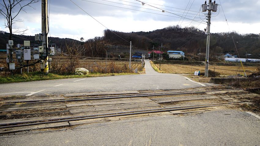 건널목에 남아 있는 옛 선로, 좌 우에 들판, 가운데 곧게 뻗은 농로, 앞을 가로 막고 있는 야산, 푸른 하늘, 우측에 전봇대에 변전 트랜스,