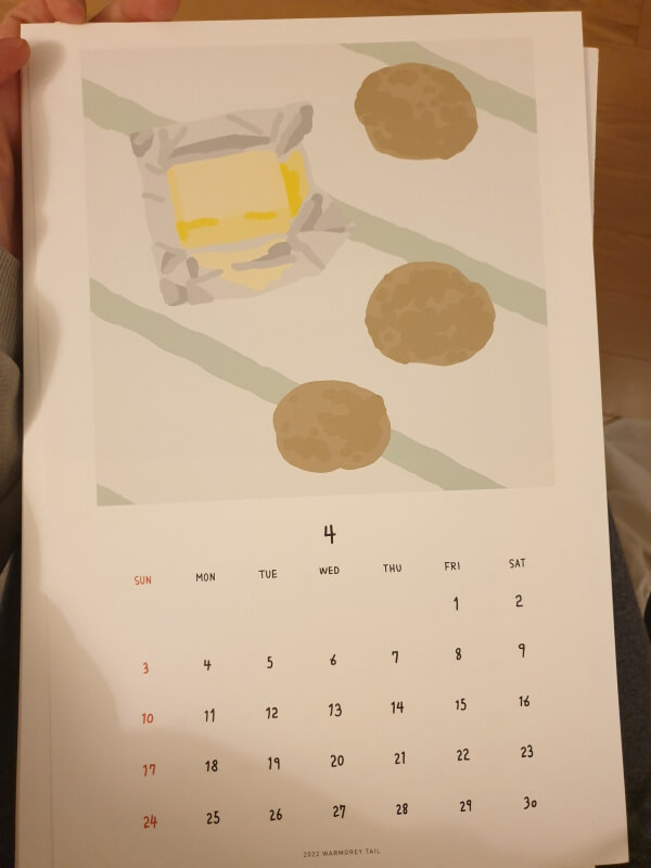 옅은 민트색의 식탁보가 깔린 식탁에 종이 포장이 벗겨진 버터와 감자 3 덩어리가 놓여진 일러스트가 그려진 4월의 웜그레이테일 달력.