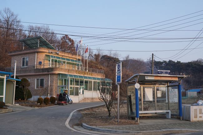 깃발이 게양된 마을회관 건물, 그 앞 길 건너에 버스정류장, 푸른 하늘,