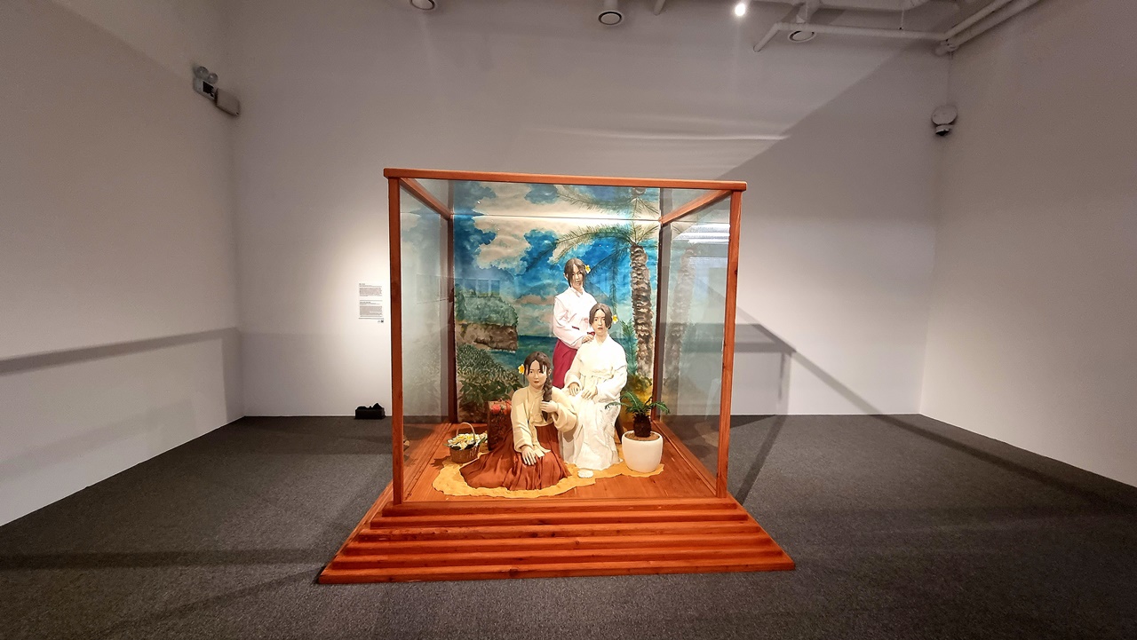 유리상자 안에 조선인 어린신부의 모습을 한 3명의 인형 작품. 주황색과 빨간색의 한복 치마와 흰색의 저고리를 입고 있는 모습