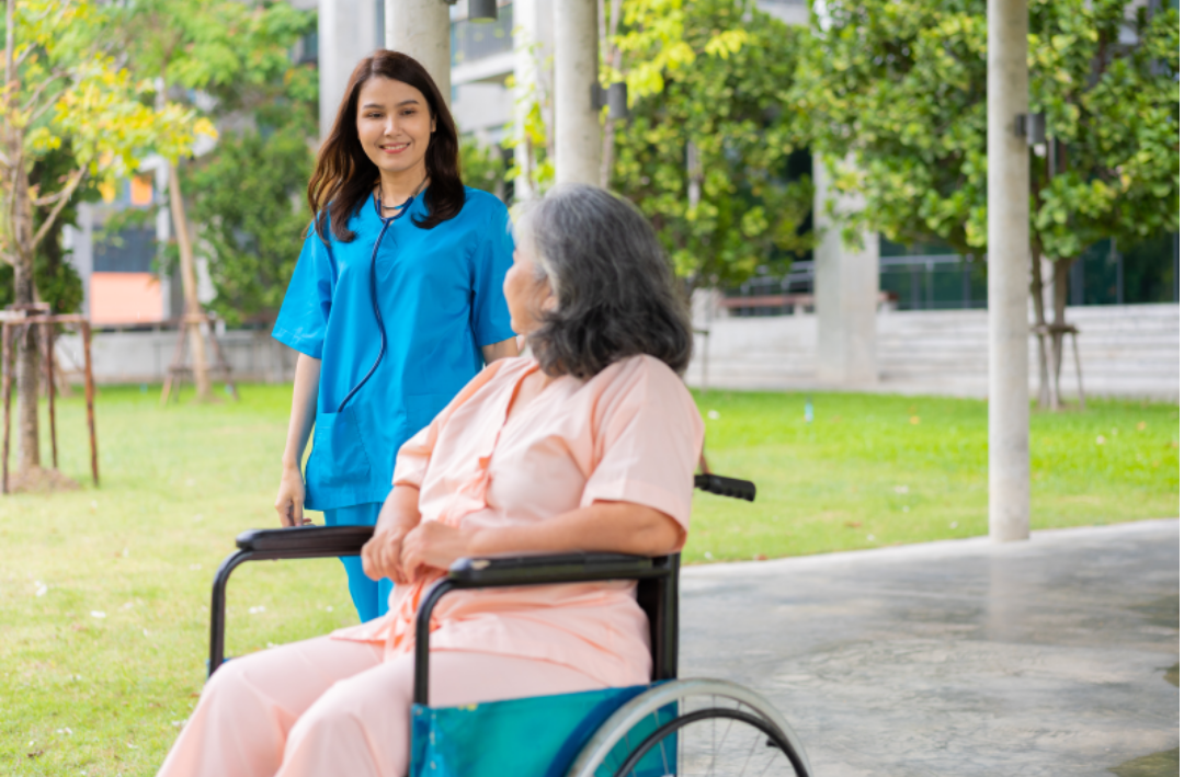 한 할머니가 휠체어에 앉아있고 간병인이 도와주고 있다.