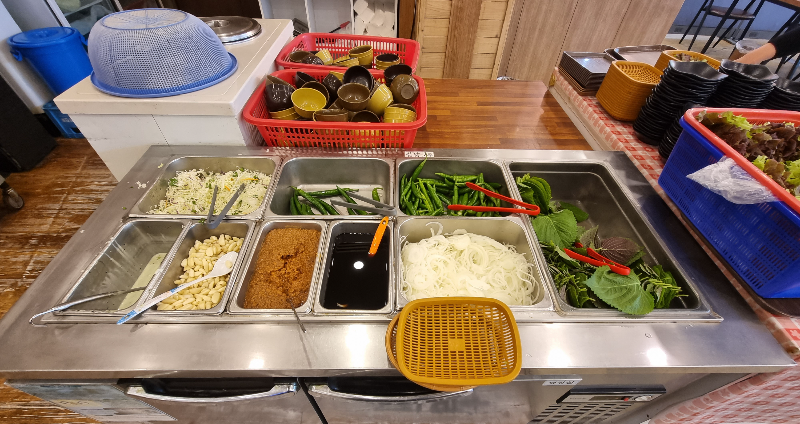 스테인리스로 된 반찬 냉장고 위에 각종 채소들이 배치되어 있는 셀프바 모습. 마늘 쌈장 고추 양파 깻잎 상추 등이 준비되어있음.