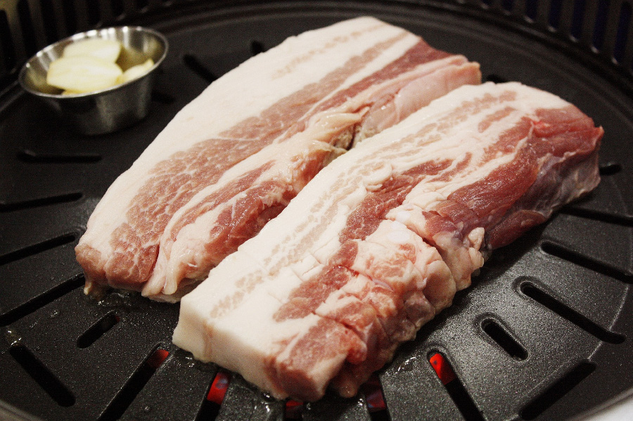 검고 둥근 철판 위에 돼지고기 삼겹살 2개의 덩이와 동그랗고 쇠로 된 작은 그릇에 마늘을 썰어 놓은 것을 놓고 같이 굽고 있는 사진