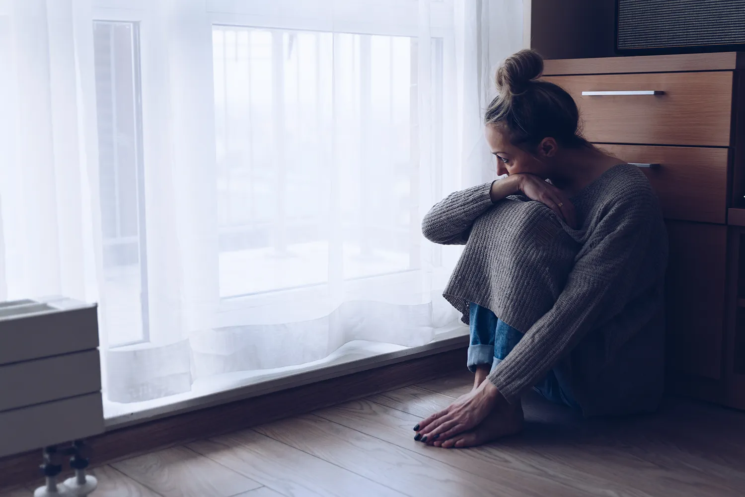 여성이 거실 바닥에 앉아 창문을 바라보고 있는 우울한 여성