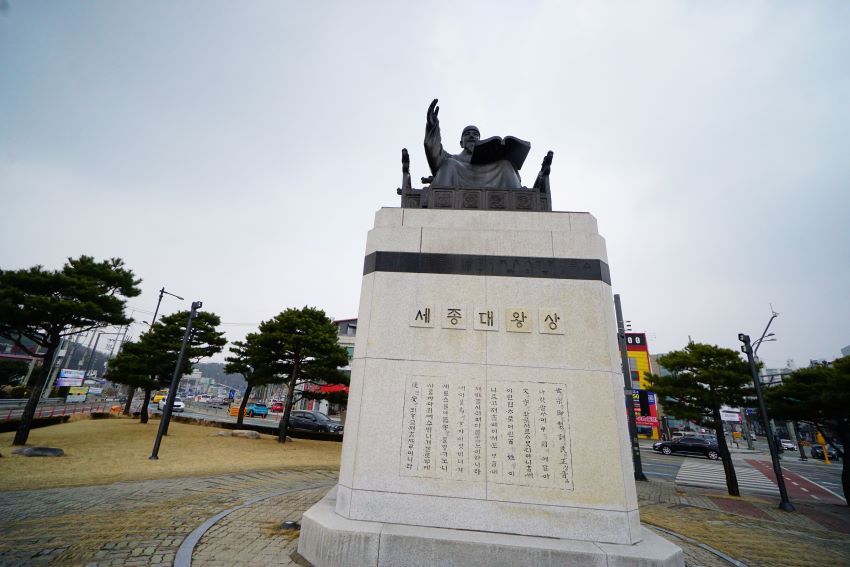 흰 대리석 사각 바탕 위에 높게 모신 세종대왕상, 올려다 본 사진, 흐린 하늘, 뒤로는 소나무들과 도시 건물들,