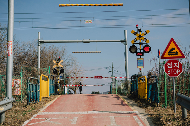 좁은 길 앞에 철도 건널목이 있다. 차단기가 내려와 있으며, 오른쪽에는 정지(Stop) 표지판도 있다.