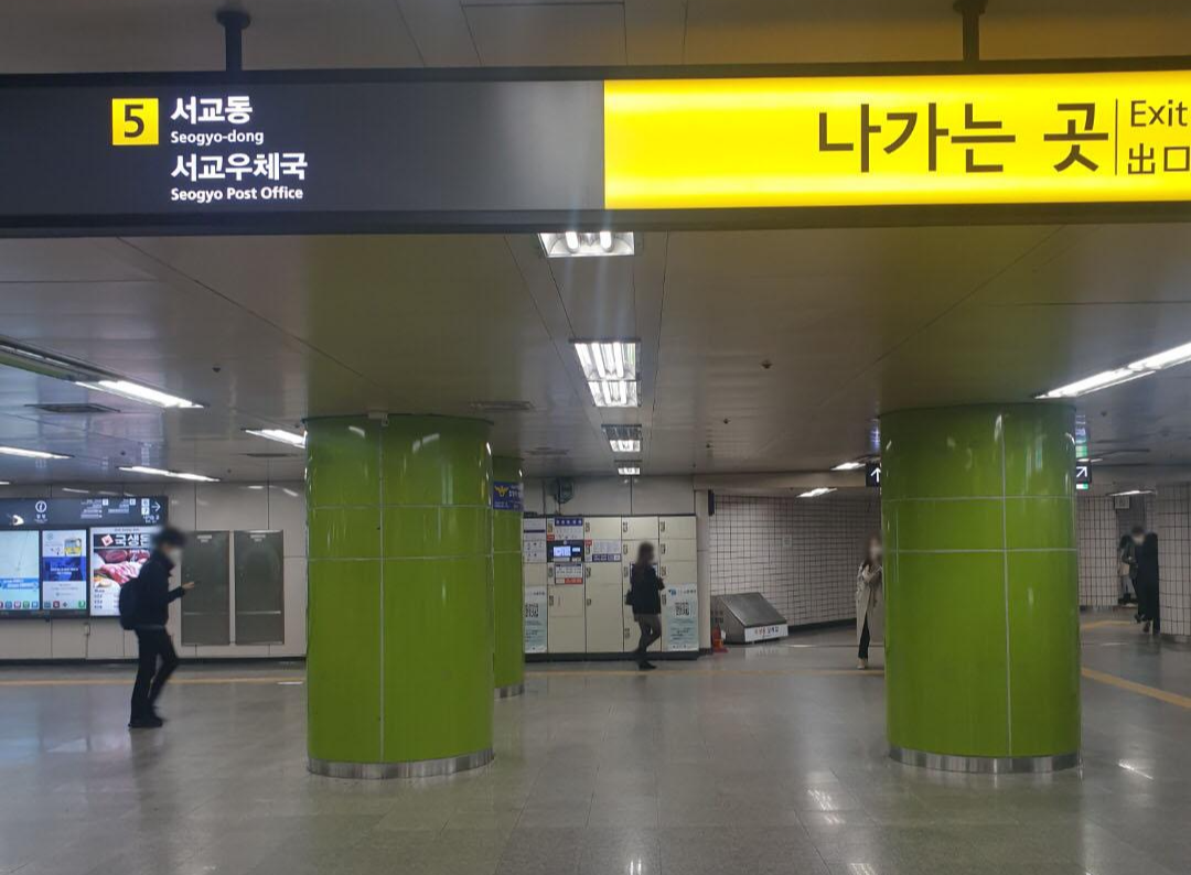 초록 기둥들이 서있고 사람들이 지나다니고 벽면에 물품보관함이 있는 지하철 역사 내 공간