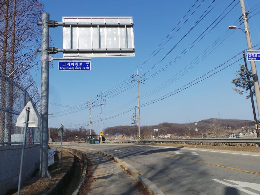 푸른 하늘&#44; 올려다 본 사진&#44; 교통표지판에 붙은 고려왕릉로 표지판&#44; 진한 파란 바탕에 흰글씨&#44; 우측에 전못데&#44; 전깃줄&#44; 좌측에 군부태 철조망&#44;