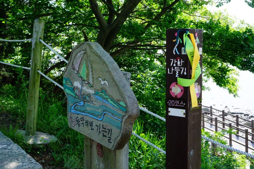 강화나들길 이정표, 검은 바탕 말뚝에 흰글씨, 이정표에 매어 놓은 길안내 리본, 왼쪽에는 목판 안내판, 동막해변 가는 길이라고 쓰여 있음, 뒤로는 큰 나무, 나무 사이 아래로 보이는 해변 모래사장,