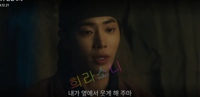 천승휘가 검은 두건을 쓰고 어두운 배경 속에서 다소 슬픈 표정으로 상대에게 이야기하고 있다. 화면 하단 자막에는 &amp;quot;내가 옆에서 웃게 해 주마&amp;quot;라는 문구가 표시되어 있다. 