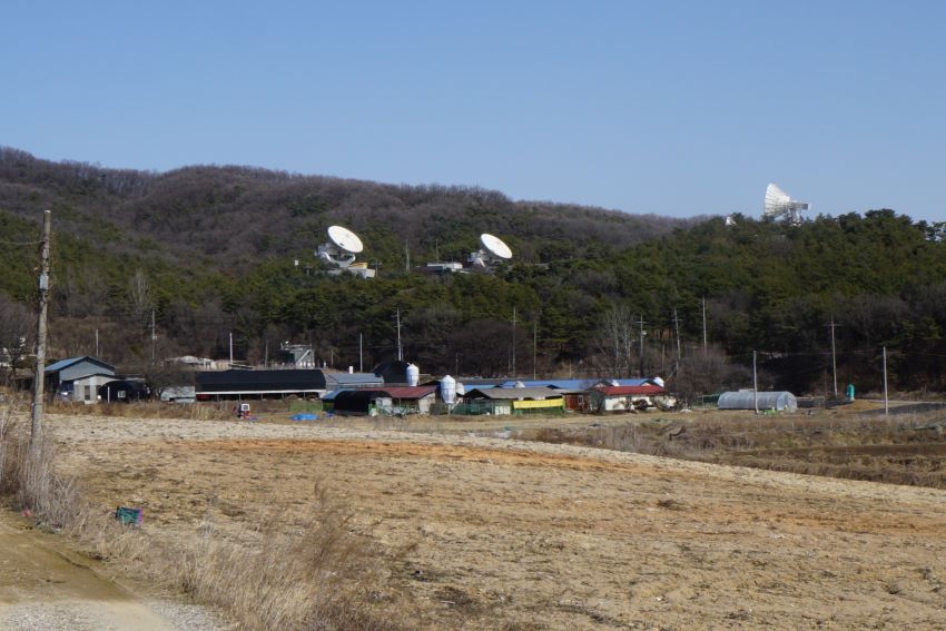 푸른 하늘&#44; 산 중턱에 네 개의 원형 대형 안테나&#44; 흰색&#44; 산자락 아래에 주택 건물들&#44; 앞쪽에 밭&#44;