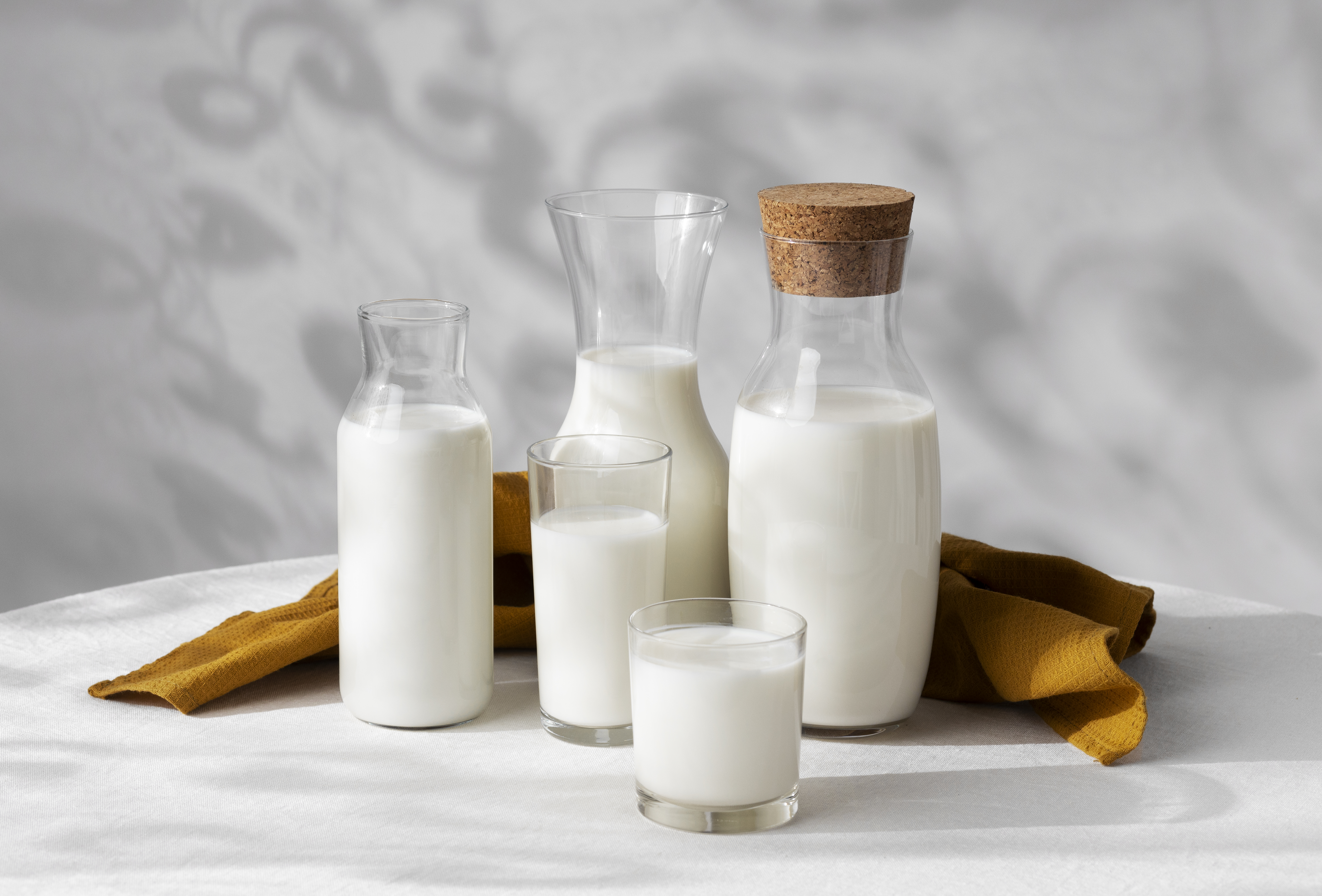 classic-glass-bottle-milk 동그랗고 옆은 회색의 테이블 위에 3개의 투명한 유리병과 2개의 투명한 유리컵에 우유를 가득 담아서 나란히 두고 그 뒤에 짙은 노란색의 천 깔판을 구겨서 놓아두고 찍은 사진