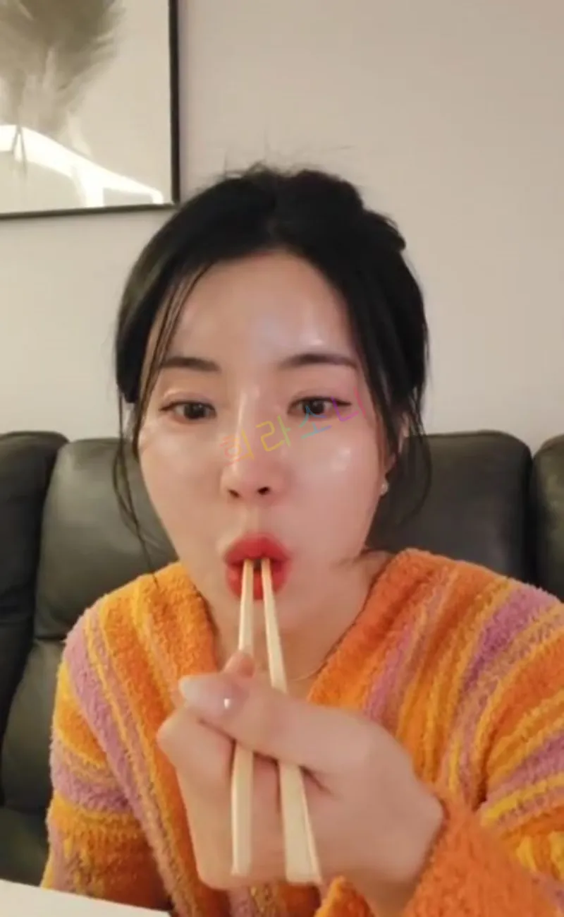 같은 여성(머리를 질끈 묶은 상태)이 젓가락 두 개를 입에 문 채 장난스러운 표정을 짓고 있다. 오렌지와 핑크 스트라이프 패턴의 가운을 입고 있으며&amp;#44; 진주 귀걸이를 착용하고 있다. 배경에는 검은 가죽 소파가 보인다. 자막에는 &amp;quot;희라소니&amp;quot;라는 텍스트가 보인다.