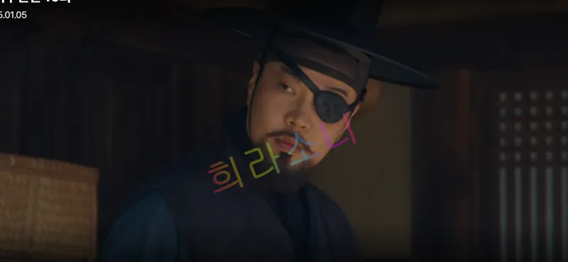 검은색 갓을 쓰고 안대를 착용한 지행수가 무표정하게 서 있습니다. 그의 시선은 옆을 향해 있으며&amp;#44; 배경은 전통적인 한옥 내부입니다.
