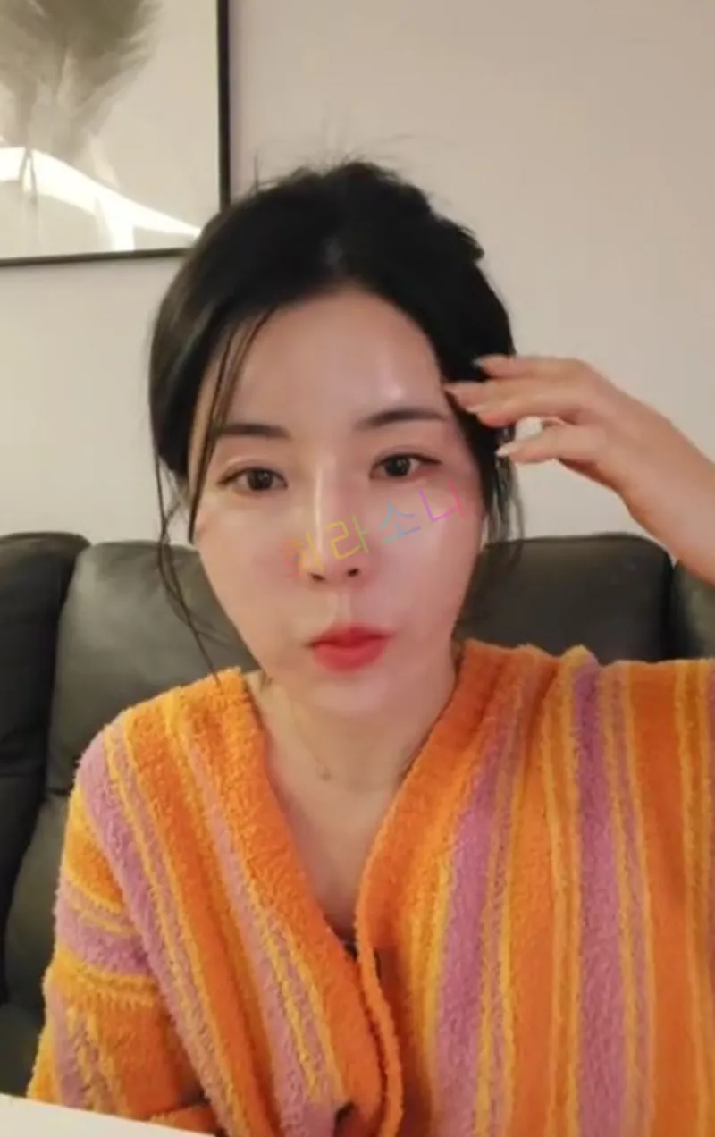 같은 여성이 한 손으로 머리를 넘기며 입을 오물거리며 음식을 먹고 있는 듯한 표정을 짓고 있다. 오렌지와 핑크 스트라이프 패턴의 가운을 입고 있으며&amp;#44; 진주 귀걸이를 착용하고 있다. 배경에는 검은 가죽 소파가 보인다. 자막에는 &amp;quot;희라소니&amp;quot;라는 텍스트가 보인다. 