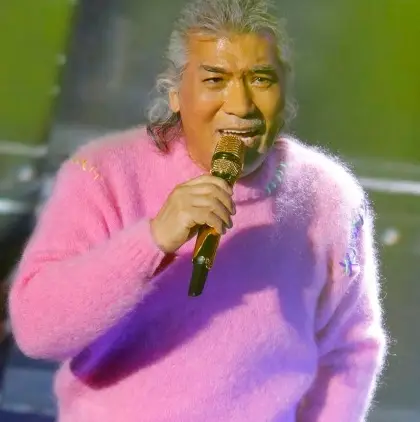 나훈아- 무대위 분홍색 니트를 입고 오른손으로 금색 마이크를 쥐고 노래하는 나훈아