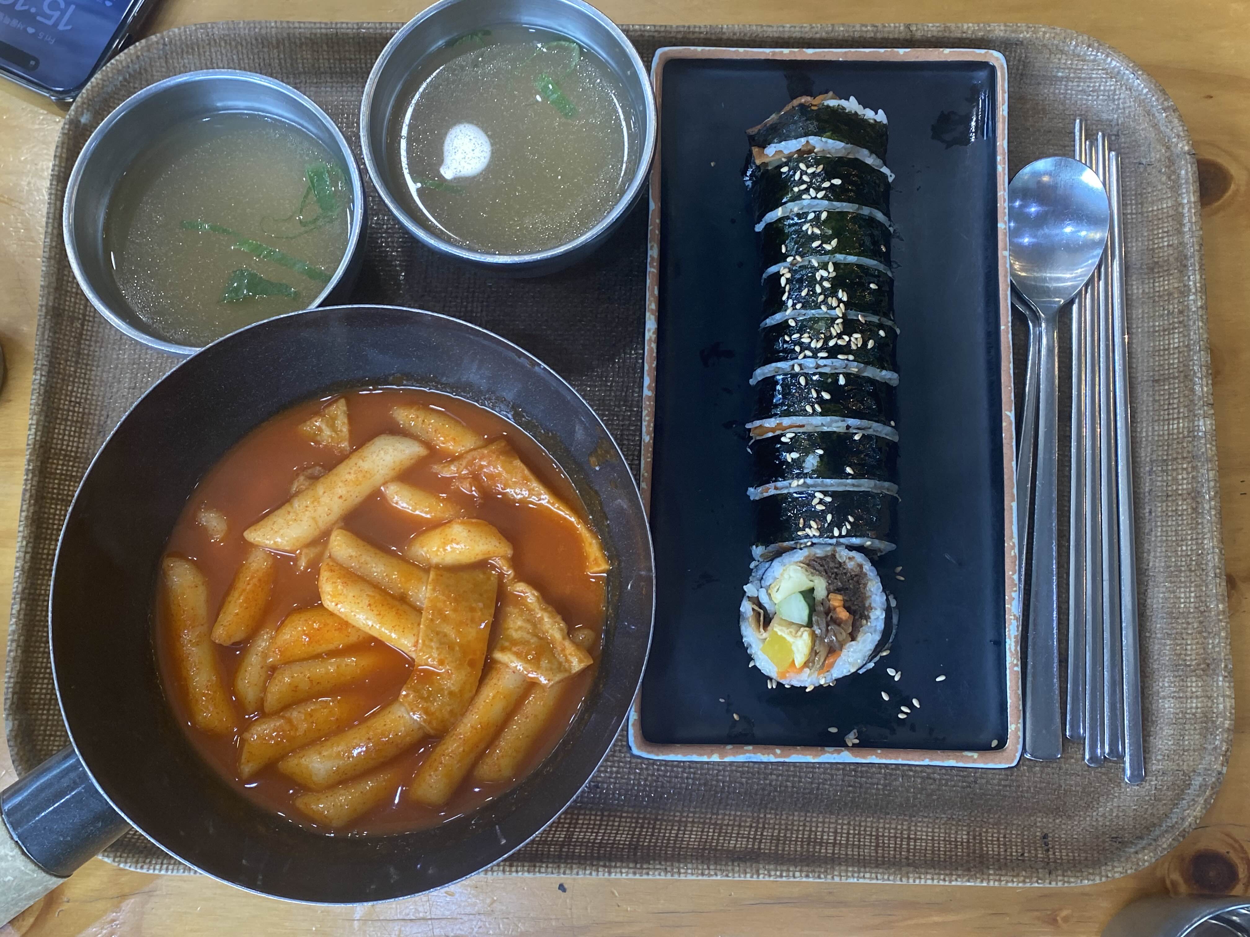 일정한 간격으로 썰린 후 깨가 뿌려진 김밥과 그 옆에 놓인 빨간 국물의 떡볶이, 그리고 두 잔의 국물